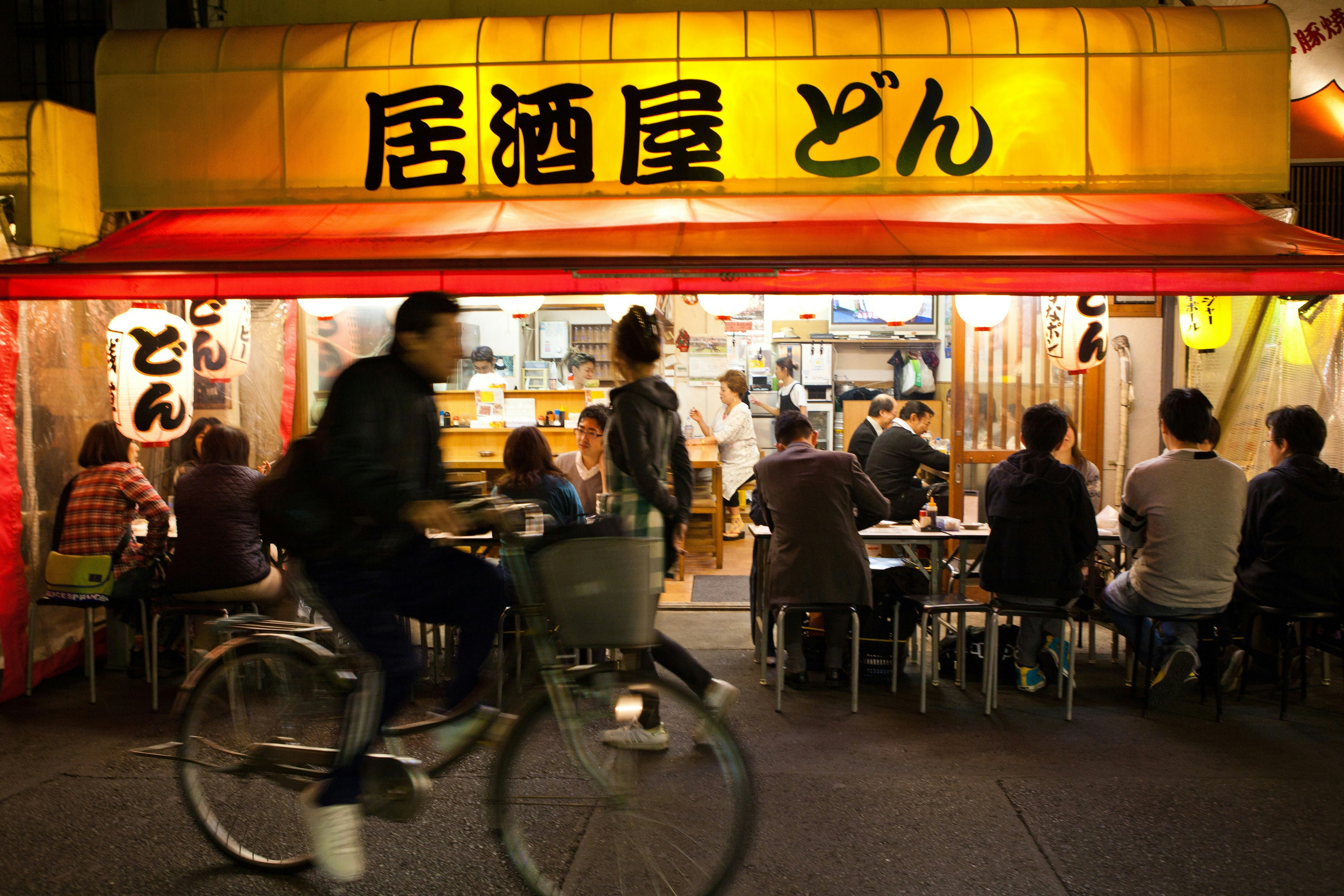 A man cycles past a cafe in Tokyo, which is open to the street, with diners and drinkers visible beneath its canopy roof and hanging lanterns